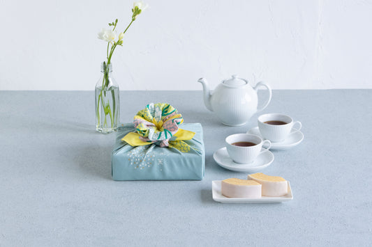 A tea time arrangement featuring a square gift box wrapped in a light green floral furoshiki cloth with a decorative flower knot, styled next to white tea cups, a teapot, and baumkuchen cake.