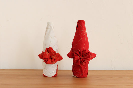 Traditional Japanese furoshiki bottle wrapping style featuring two wine bottles beautifully wrapped in red and white geometric patterned cloths with decorative floral knots on a wooden table.