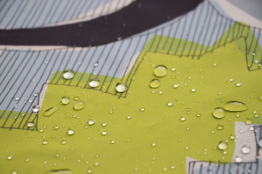 A detailed close-up showing water droplets sitting on a water-resistant fabric. The fabric features a modern illustration with bright green shapes, fine vertical black lines, and a light blue background.