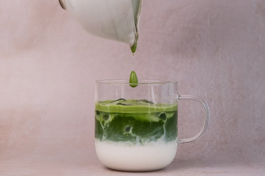 Layered iced matcha latte with milk and ceremonial matcha, pouring matcha from matcha bowl into glass mug on pastel pink background.