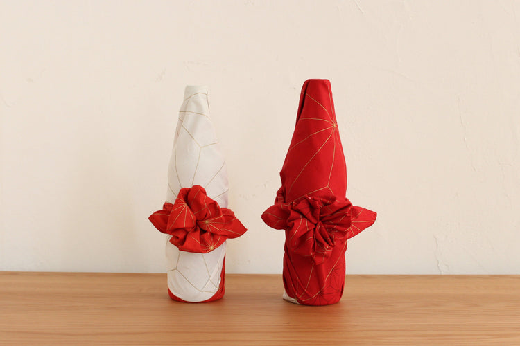 Traditional Japanese furoshiki bottle wrapping style featuring two wine bottles beautifully wrapped in red and white geometric patterned cloths with decorative floral knots on a wooden table.