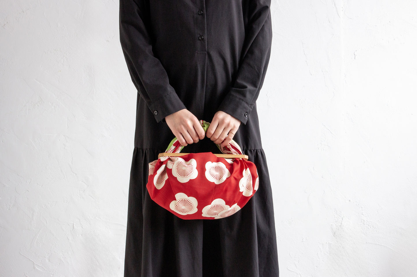 A person in a minimalist black dress holding a DIY handmade handbag created from a red and white apricot blossom pattern Japanese furoshiki cloth attached to a wooden handle.