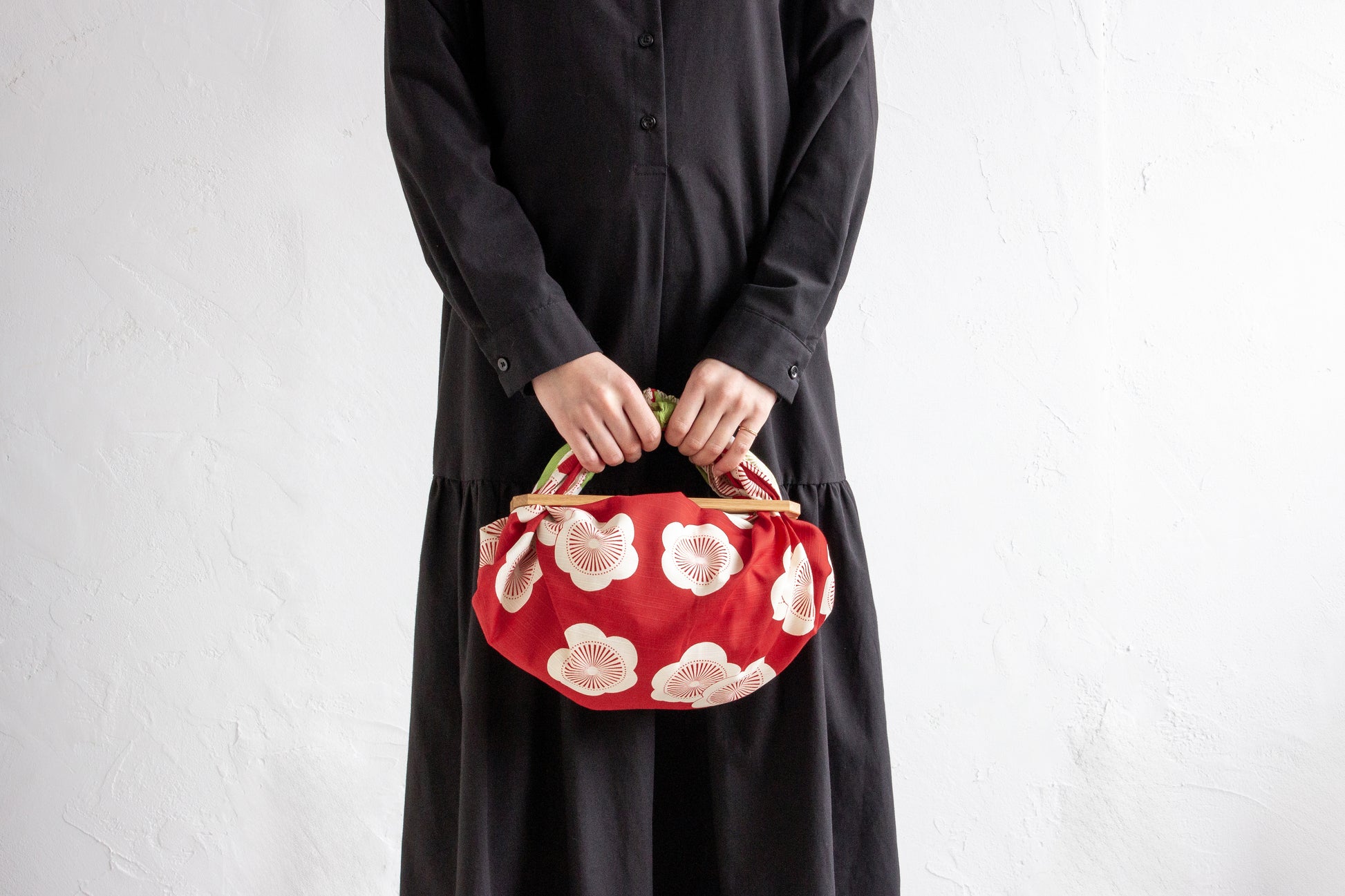 A person in a minimalist black dress holding a DIY handmade handbag created from a red and white apricot blossom pattern Japanese furoshiki cloth attached to a wooden handle.