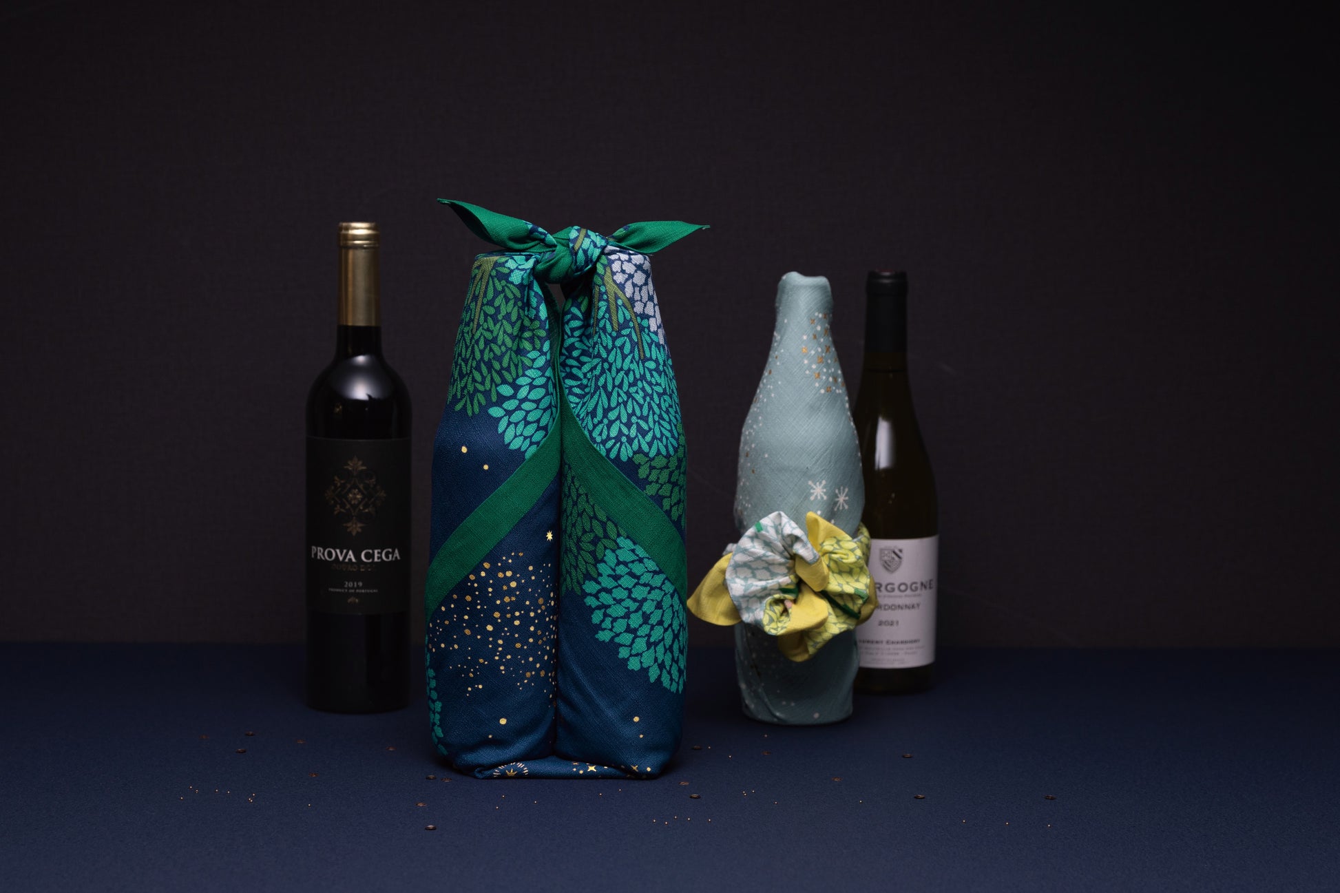 Two wine bottles beautifully wrapped in decorative Japanese furoshiki cloths using a two-bottle knot technique, styled against a dark minimalist background.
