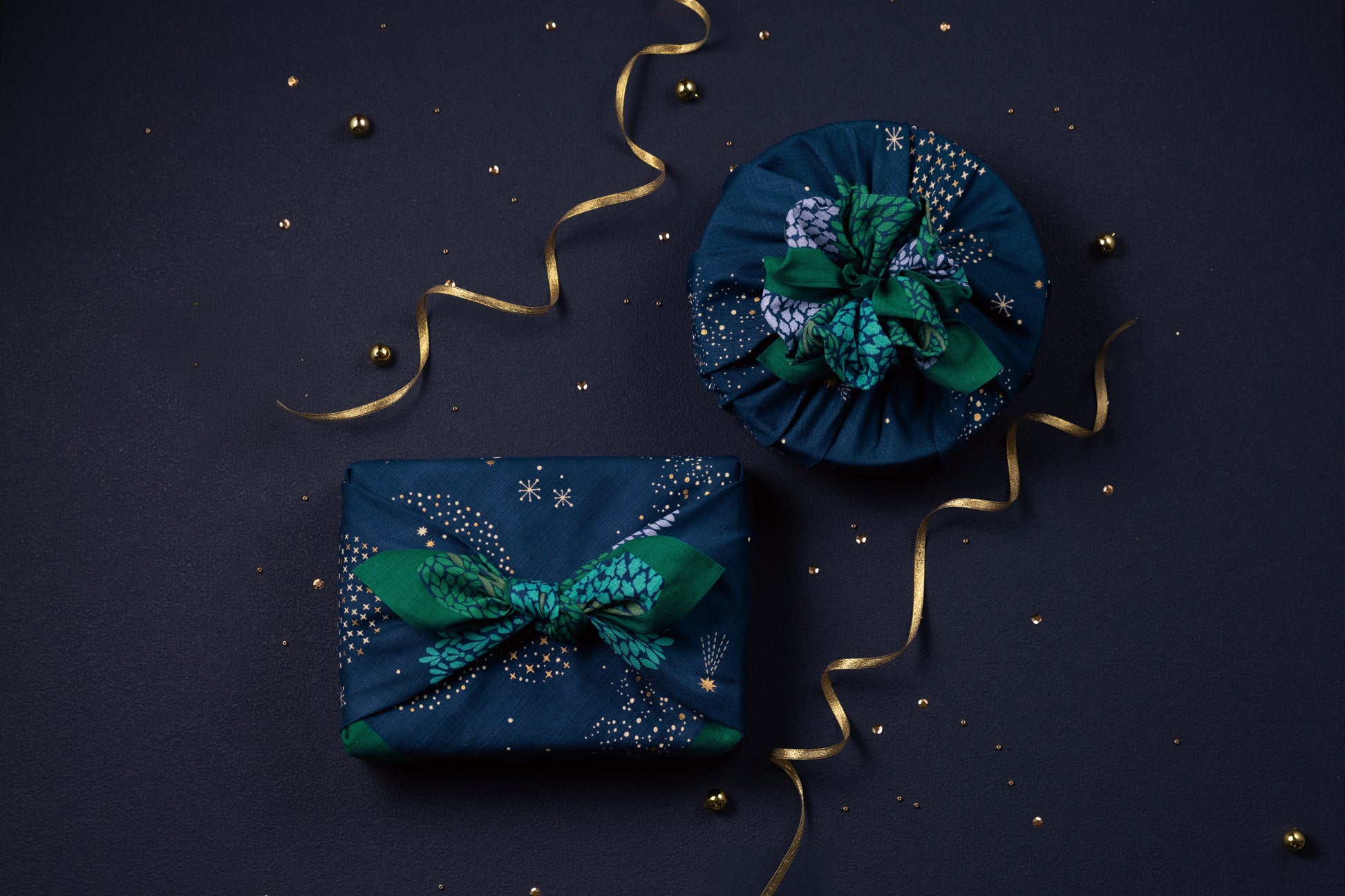 Overhead shot of square and round gift boxes wrapped in a navy blue starry night furoshiki cloth, decorated with elegant gold ribbons and matching confetti sparkle.