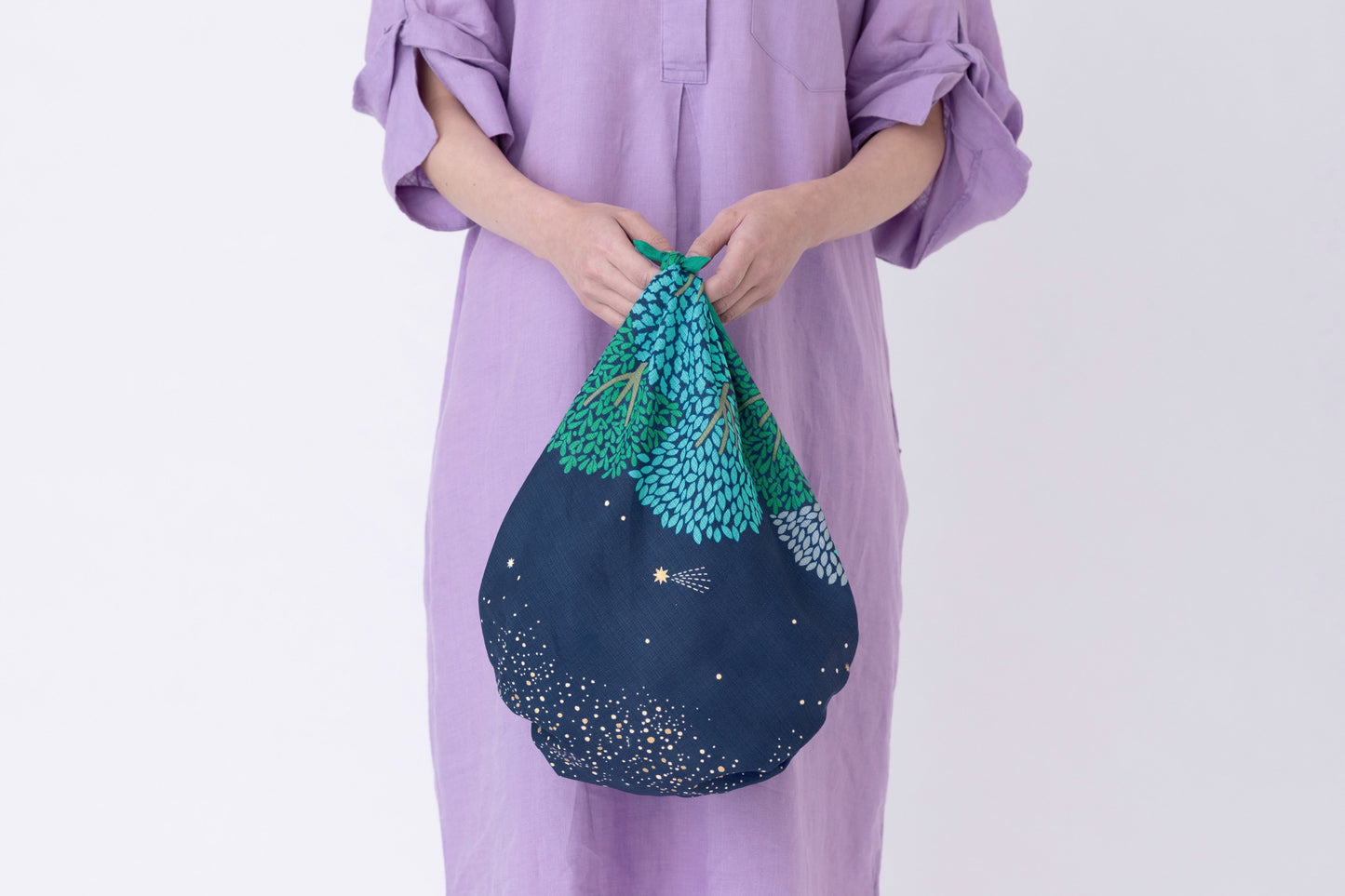 A person wearing a lilac purple linen tunic dress holding a slouchy handbag tied from a dark blue and green tree pattern Japanese furoshiki fabric.
