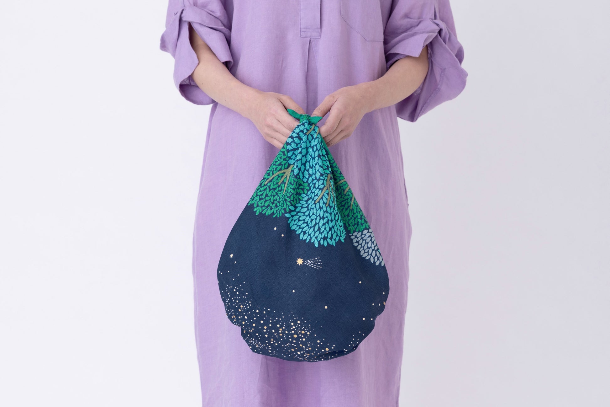 A person wearing a lilac purple linen tunic dress holding a slouchy handbag tied from a dark blue and green tree pattern Japanese furoshiki fabric.