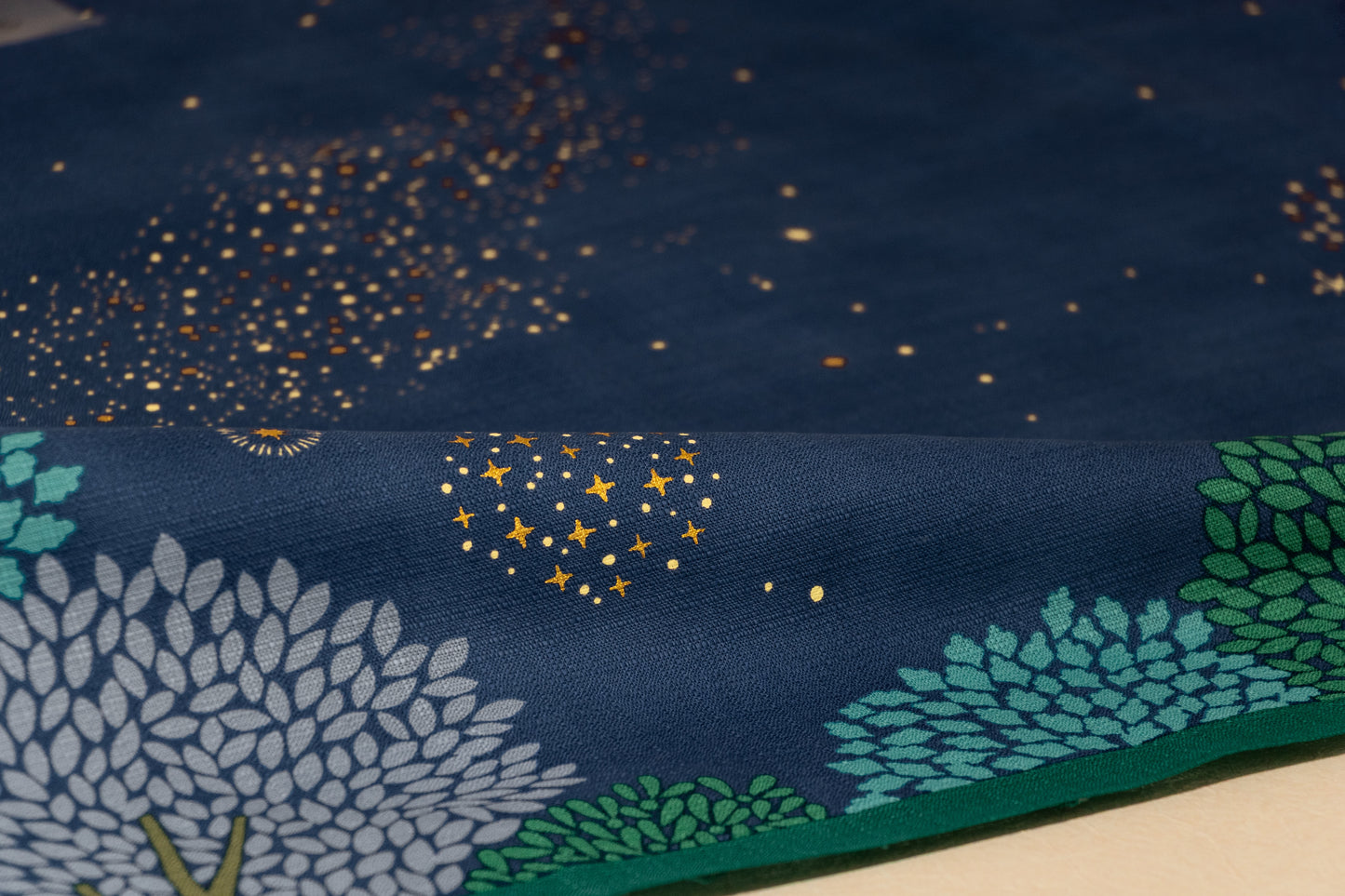 Close-up of the woven cotton texture of a Japanese furoshiki fabric, highlighting detailed gold metallic ink star clusters and stylized green and blue trees.
