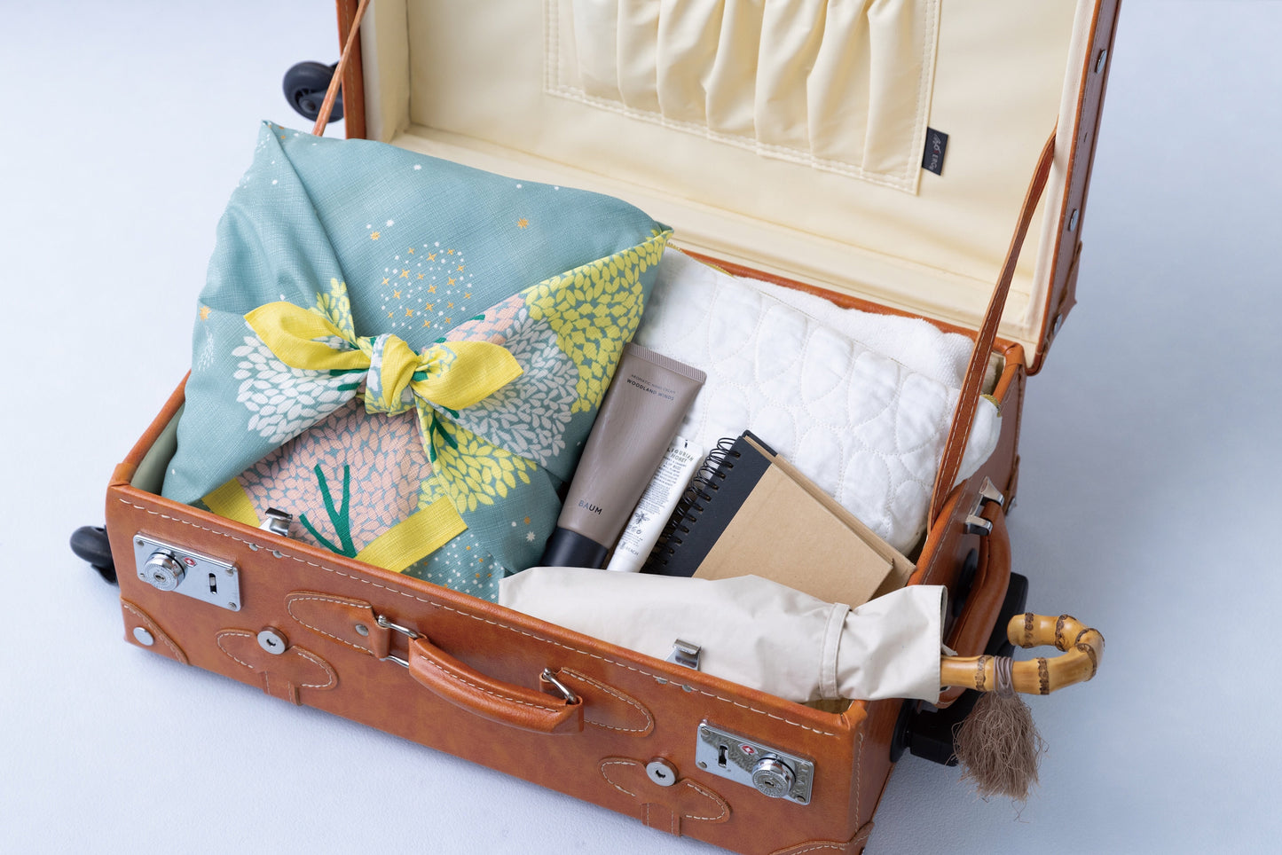 A sage green floral furoshiki cloth used as an eco-friendly travel packing organizer inside a vintage leather suitcase alongside toiletries, a notebook, and an umbrella.