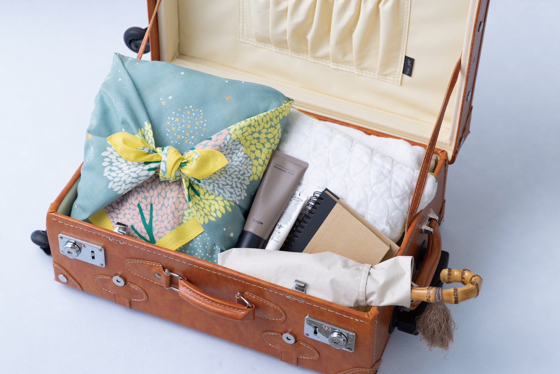 A sage green floral furoshiki cloth used as an eco-friendly travel packing organizer inside a vintage leather suitcase alongside toiletries, a notebook, and an umbrella.