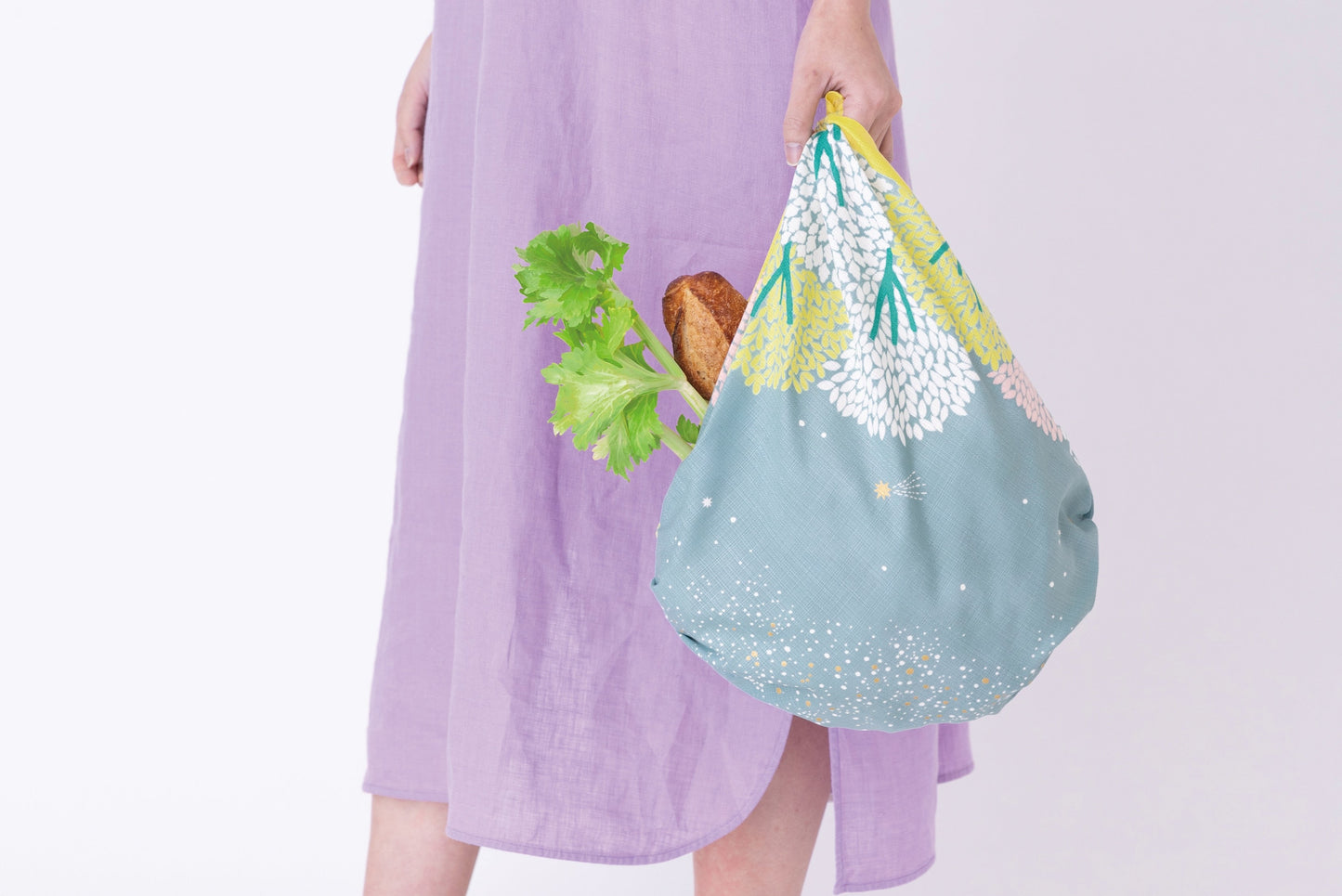 A person wearing a lavender linen dress holding an eco-friendly grocery tote bag tied from a sage green and yellow floral Japanese furoshiki cloth, filled with fresh celery and bread.