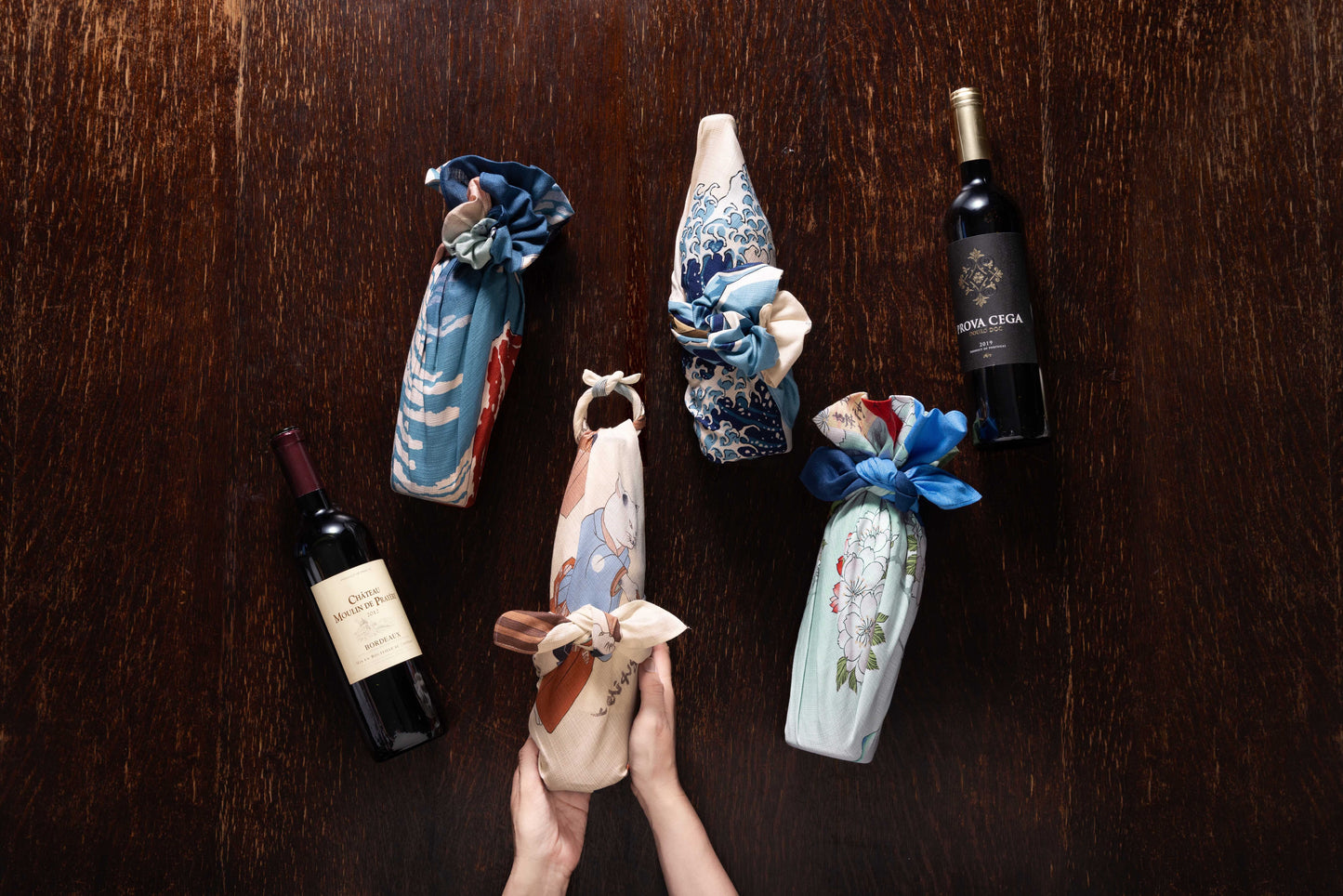 Overhead view of hands holding wine bottles decoratively wrapped in assorted traditional Japanese furoshiki cloths with ukiyo-e prints and floral motifs on a dark wood background.