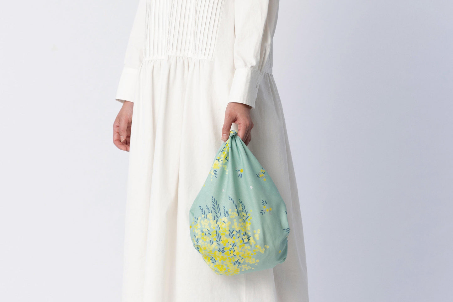 A woman in a long white pleated dress holding a reusable grocery bag knot-tied from a mint green and yellow mimosa pattern Japanese furoshiki cloth.