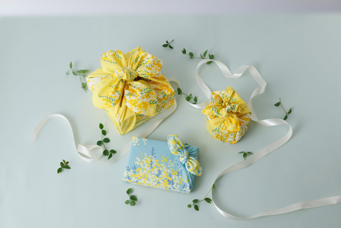 Two fabric gift pouches with drawstring closures, one in bright yellow floral print and one in light blue floral print, arranged on a pale gray background with white ribbons and small green leaves.