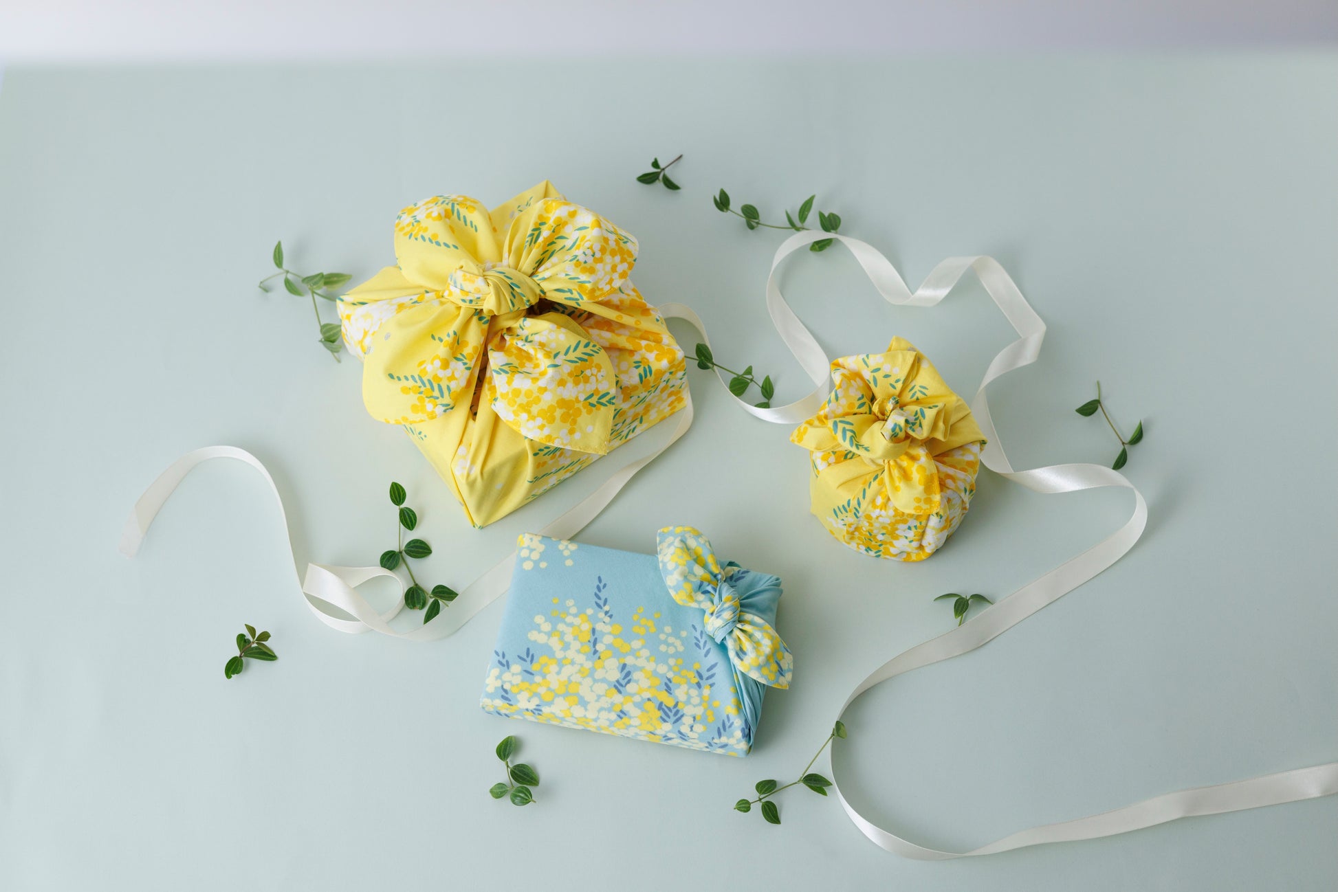 Two fabric gift pouches with drawstring closures, one in bright yellow floral print and one in light blue floral print, arranged on a pale gray background with white ribbons and small green leaves.
