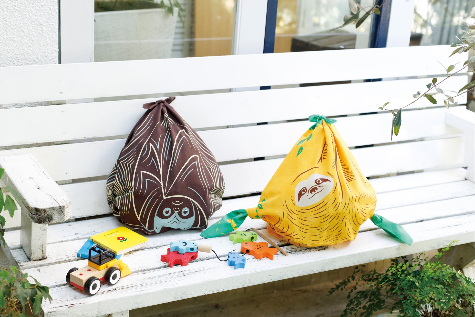 Two Japanese furoshiki cloths tied into reusable bags—one brown bat pattern and one yellow sloth pattern—resting on a white wooden garden bench next to children's wooden toys.