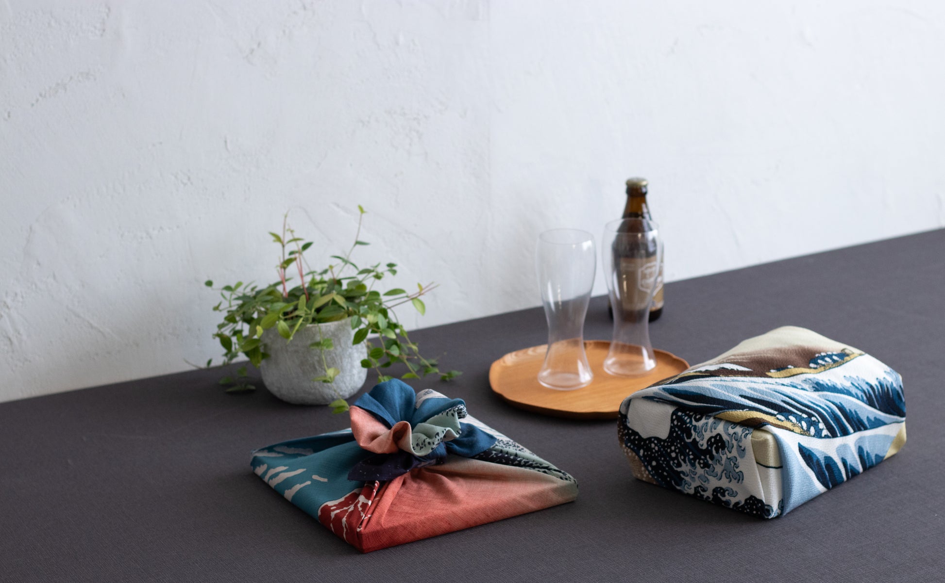Modern home styling showing a rectangular box and a flat book wrapped in Hokusai-inspired Japanese furoshiki cloth, arranged with beer glasses and a small houseplant.