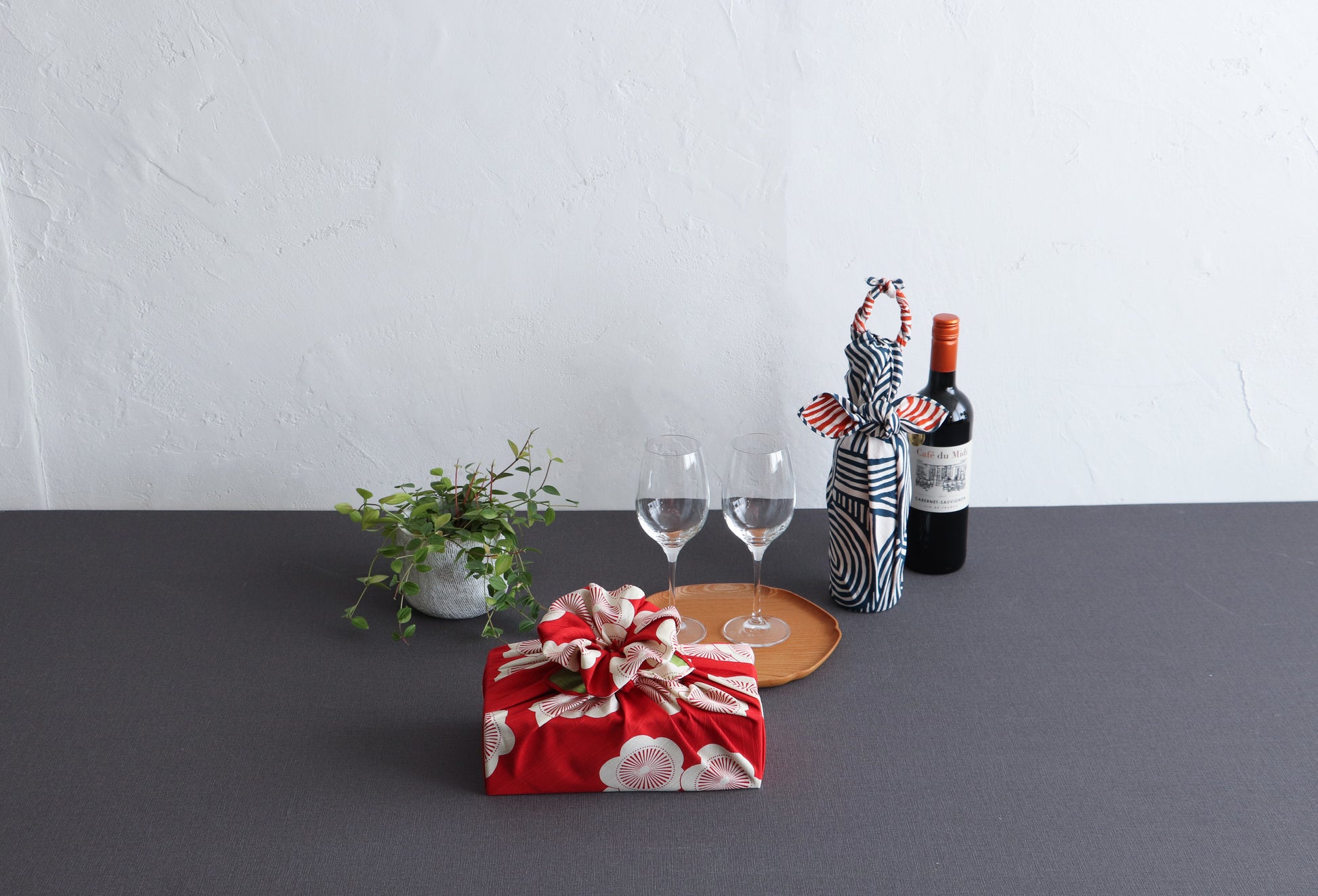 Modern table setting with a box wrapped in a red apricot blossom furoshiki and a wine bottle wrapped in a blue geometric Japanese cloth, styled with wine glasses and a small potted plant.