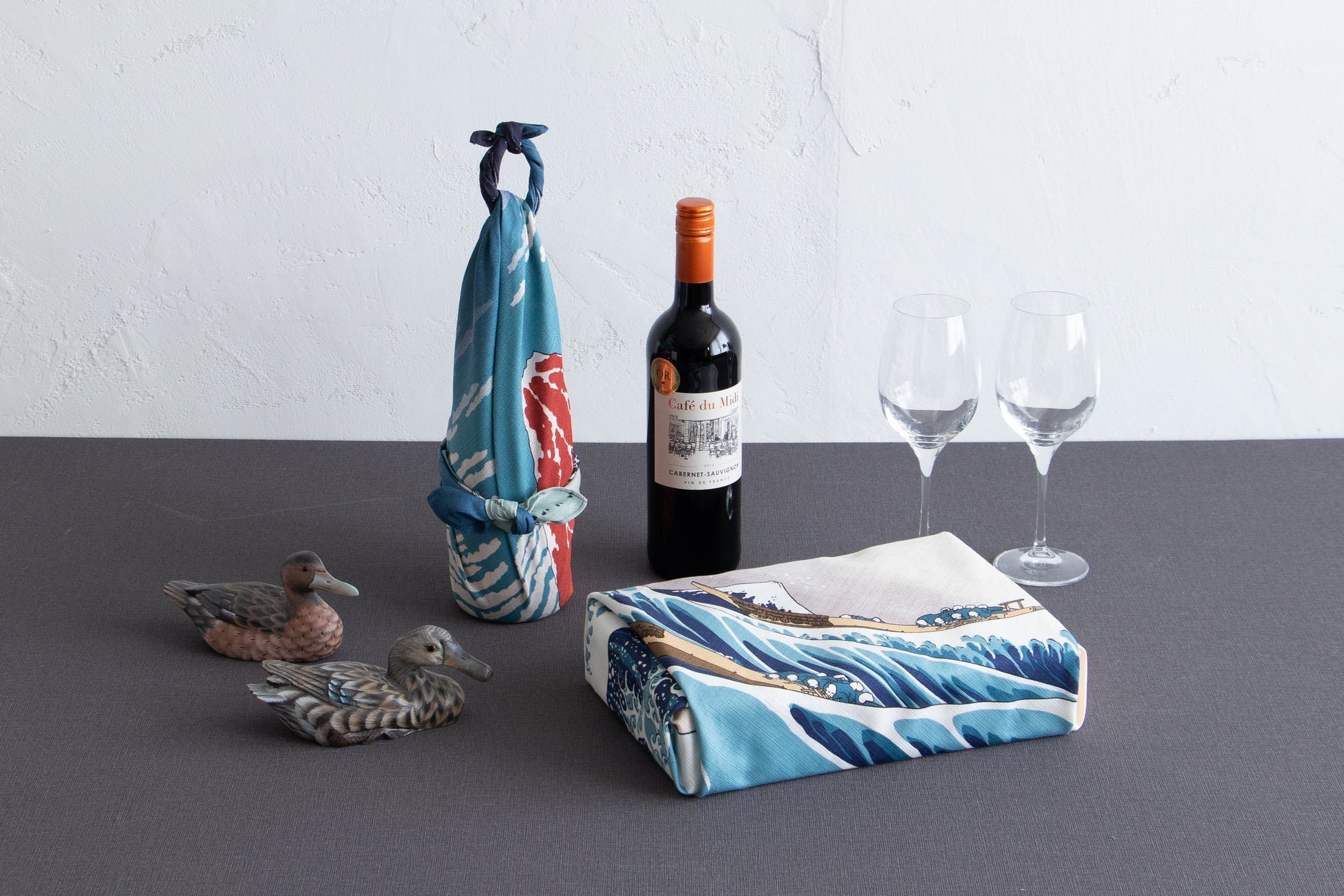 A gift box and wine bottle elegantly wrapped in Hokusai's Great Wave pattern furoshiki cloth, styled with two wine glasses and small duck figurines on a tabletop.