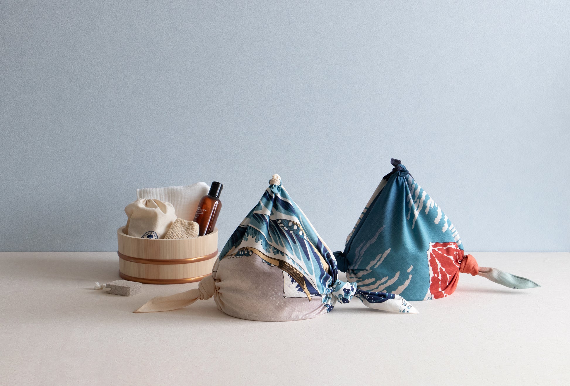Two traditional Japanese sauna or bath bags created from knot-tied furoshiki cloths featuring ukiyo-e wave art, sitting next to a wooden bath bucket with skincare amenities.