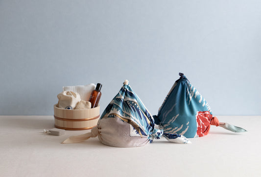 Two traditional Japanese sauna or bath bags created from knot-tied furoshiki cloths featuring ukiyo-e wave art, sitting next to a wooden bath bucket with skincare amenities.