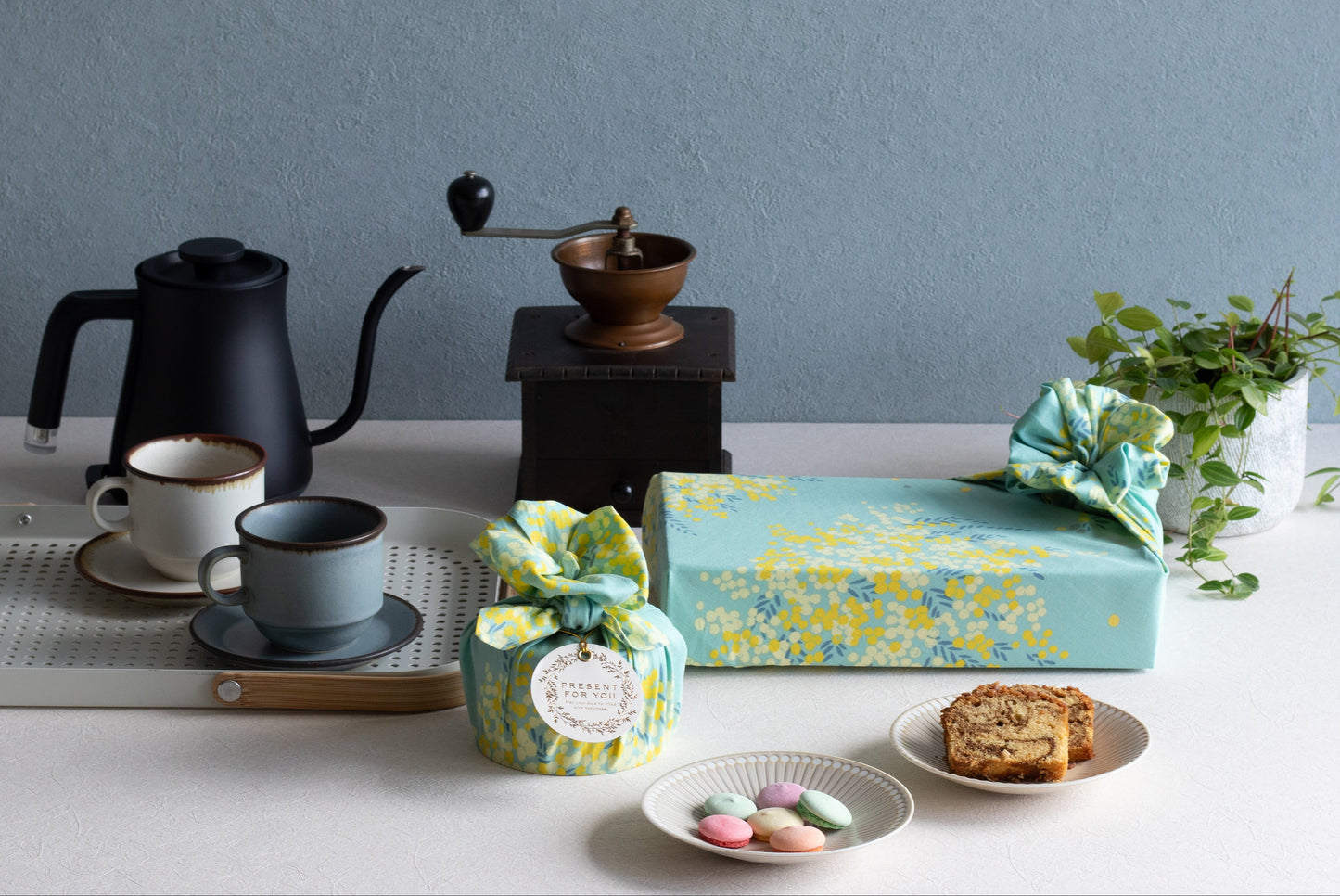 Eco-friendly gift ideas showing three packages wrapped in yellow and mint green mimosa flower Japanese furoshiki cloth, accented with white ribbon and green leaves.