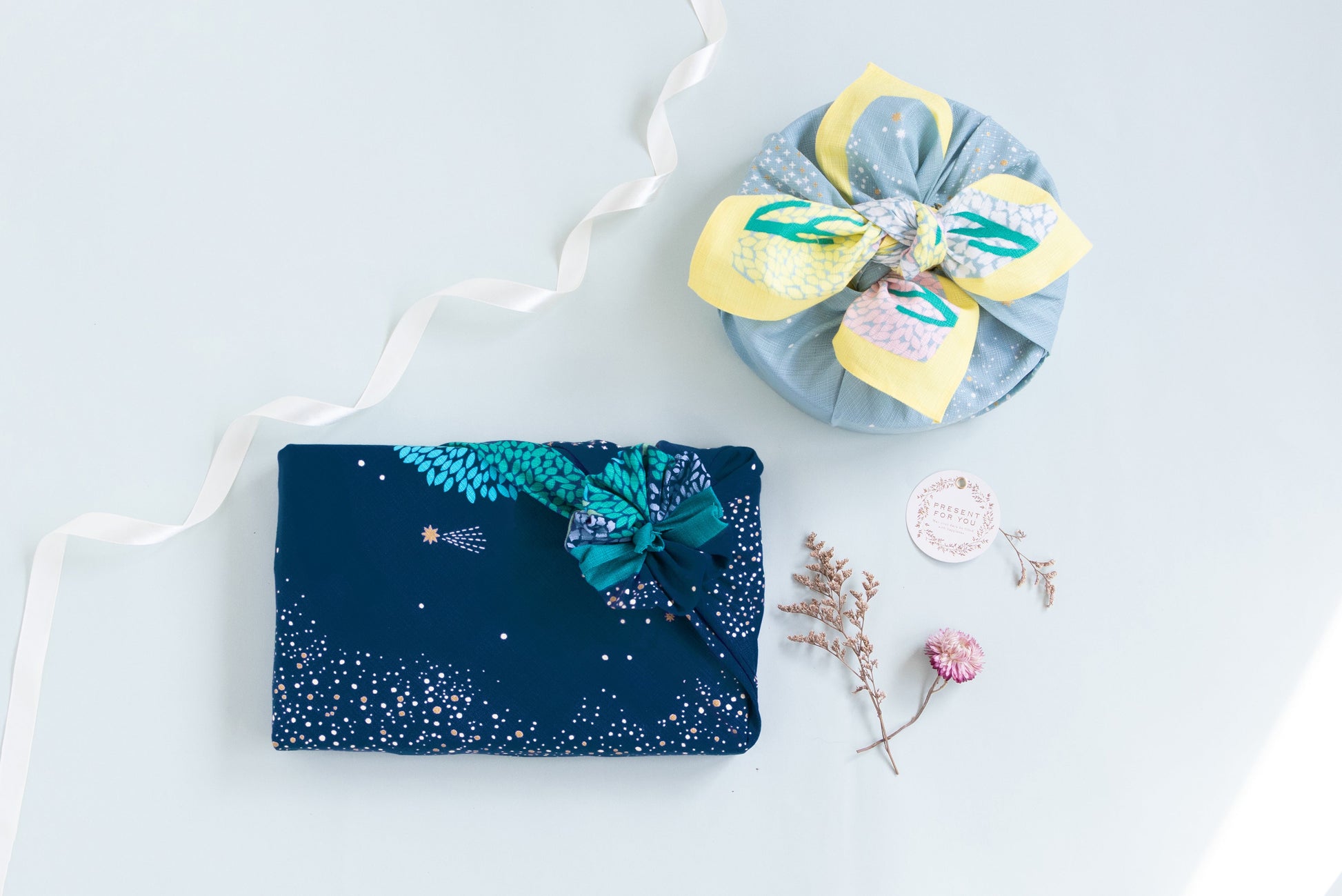 High-angle shot of two elegant Japanese gift-wrapping styles, featuring a square gift box wrapped in navy blue constellation fabric and a round gift wrapped in a light green floral furoshiki cloth.