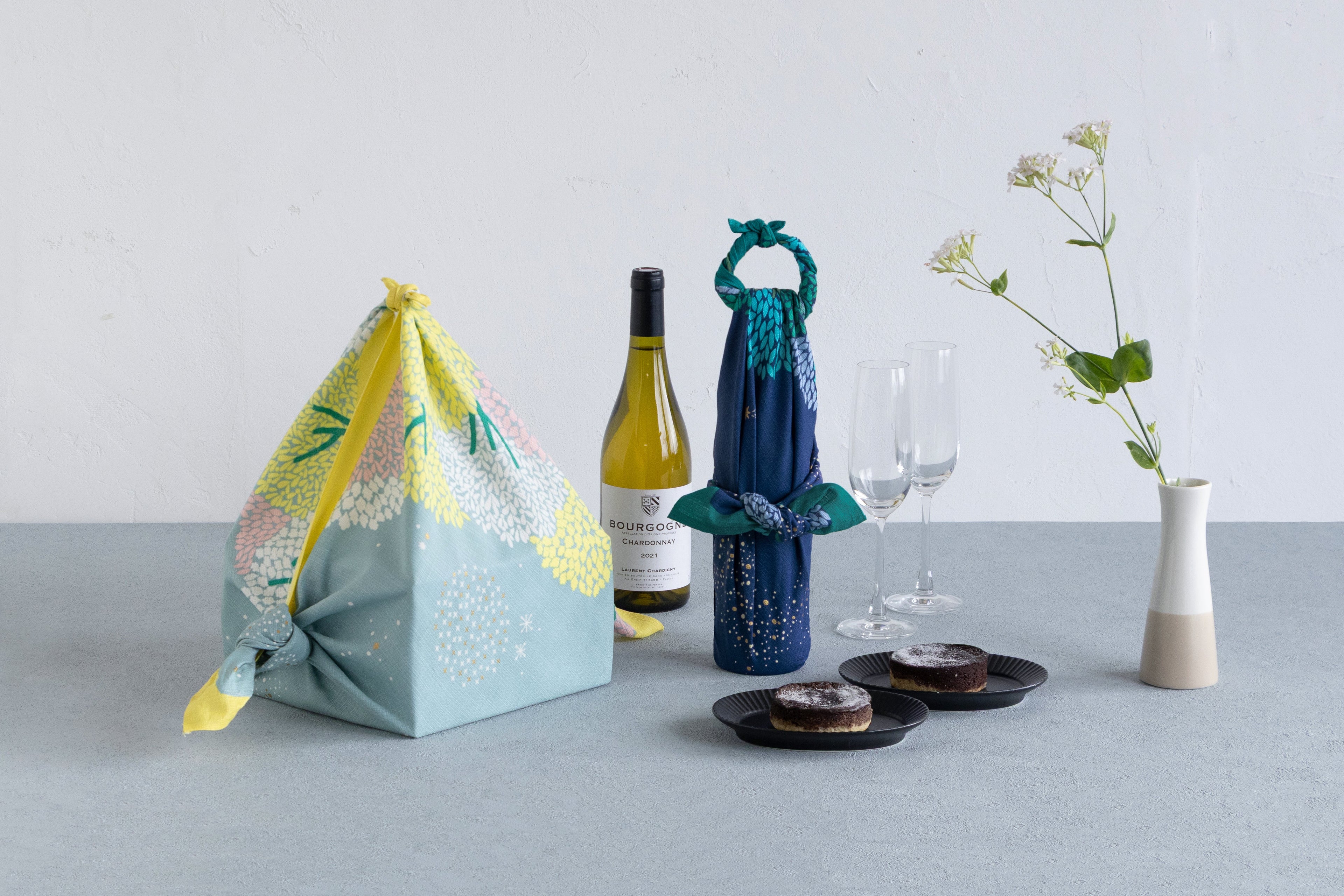 A dining tabletop setting featuring a pyramid-shaped furoshiki gift bag and a wine bottle wrapped in light blue and green floral Japanese furoshiki cloths next to a wine bottle, wine glasses, and pound cake.