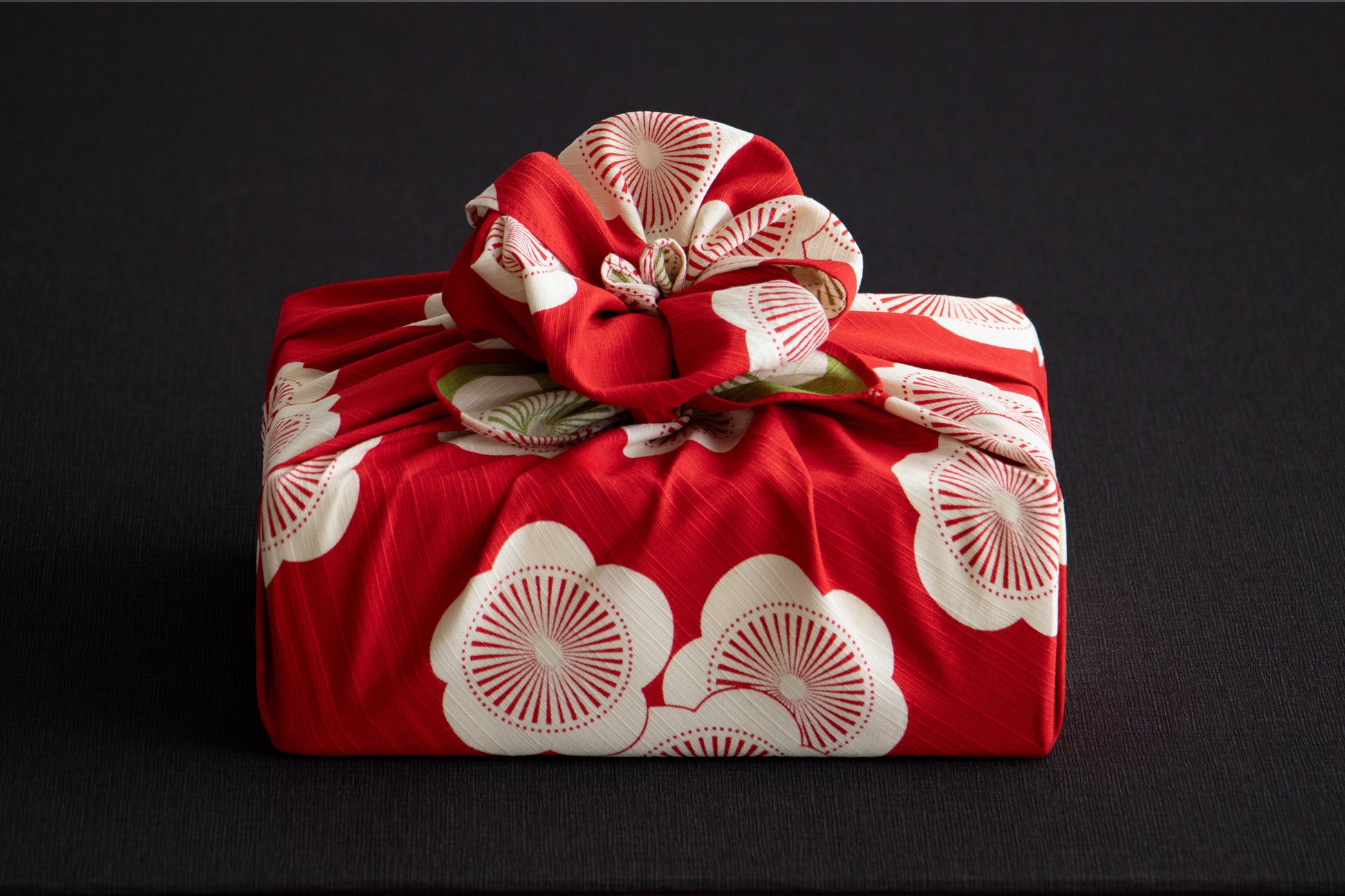 Traditional Japanese furoshiki fabric in vibrant red with a white apricot blossom pattern, elegantly wrapped around a square gift box with an intricate decorative flower knot on top.