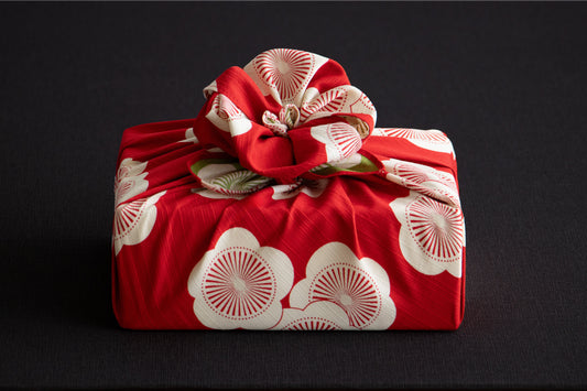 Traditional Japanese furoshiki fabric in vibrant red with a white apricot blossom pattern, elegantly wrapped around a square gift box with an intricate decorative flower knot on top.