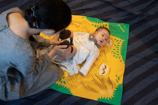 A mother using a yellow and green cartoon sloth pattern Japanese furoshiki cloth as a soft changing mat for her baby on a striped blanket.