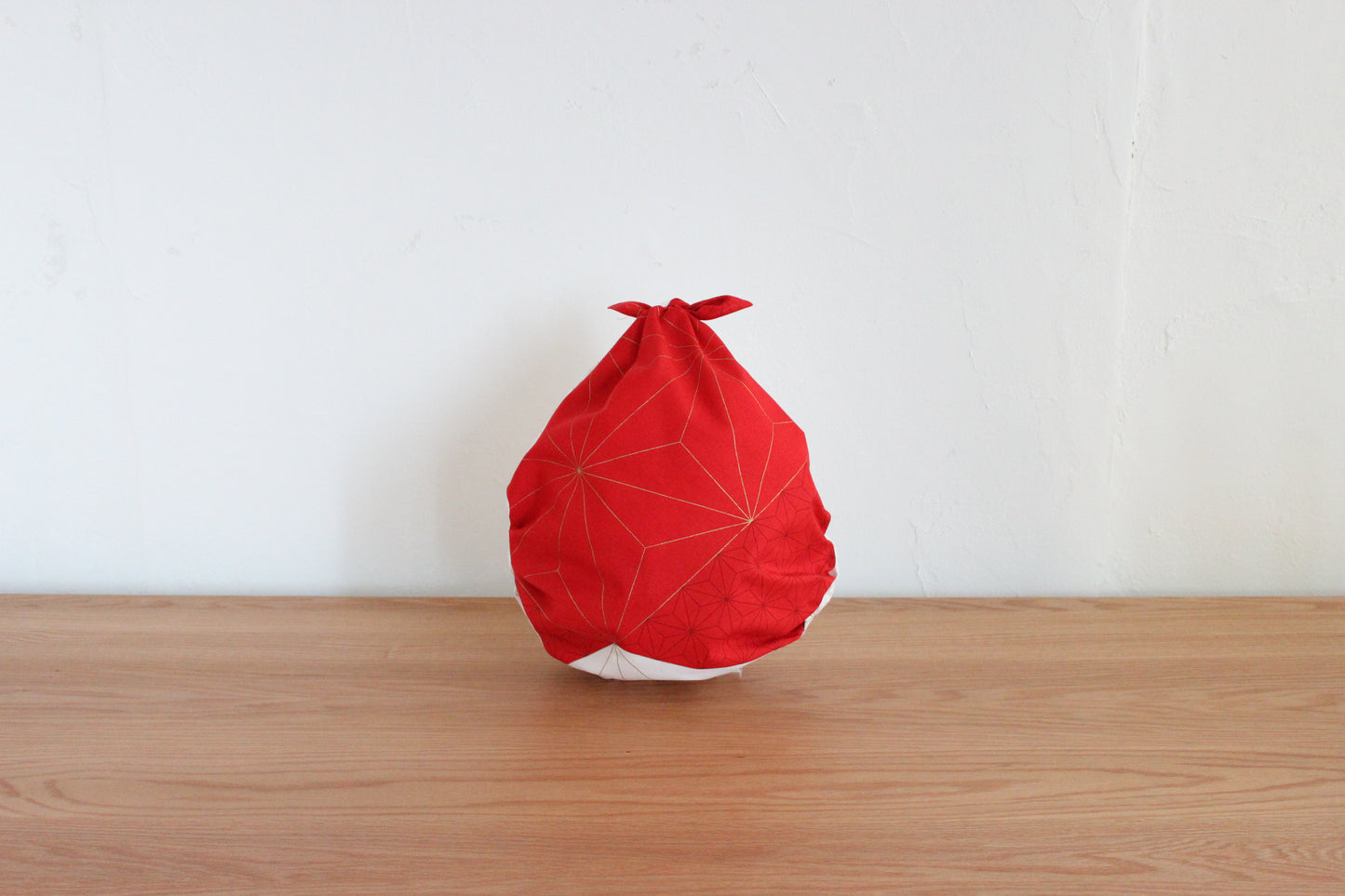 Reusable Japanese furoshiki tote bag tied into a drop shape on a wooden table, showcasing the red side of a reversible cloth with gold hemp leaf line details.