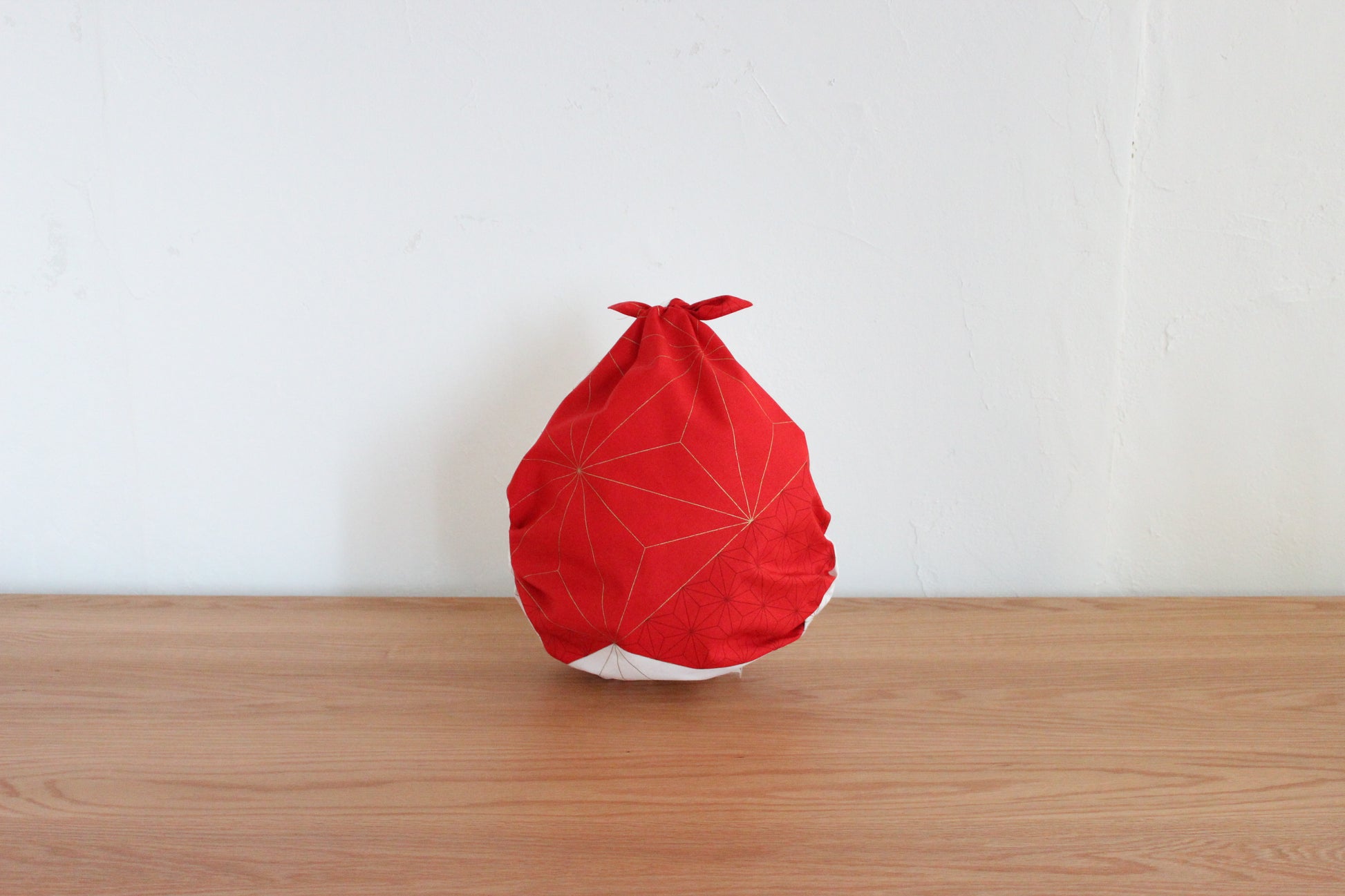 Reusable Japanese furoshiki tote bag tied into a drop shape on a wooden table, showcasing the red side of a reversible cloth with gold hemp leaf line details.
