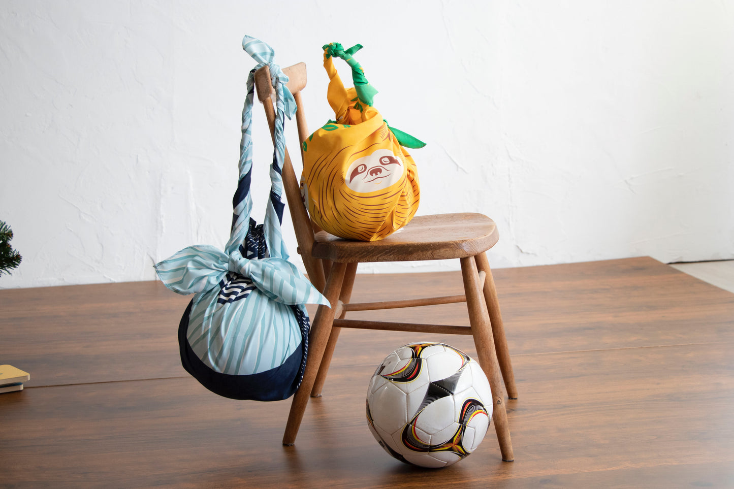 A yellow sloth-patterned furoshiki bag on a wooden chair alongside a blue striped fabric bag, next to a soccer ball on a wooden floor, demonstrating versatile wrapping.