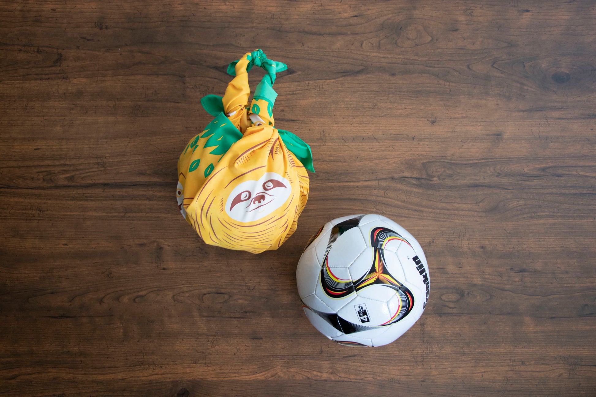Overhead view of a yellow Japanese furoshiki wrapping cloth tied into a ball-carrying bag next to a soccer ball on a dark wooden floor.