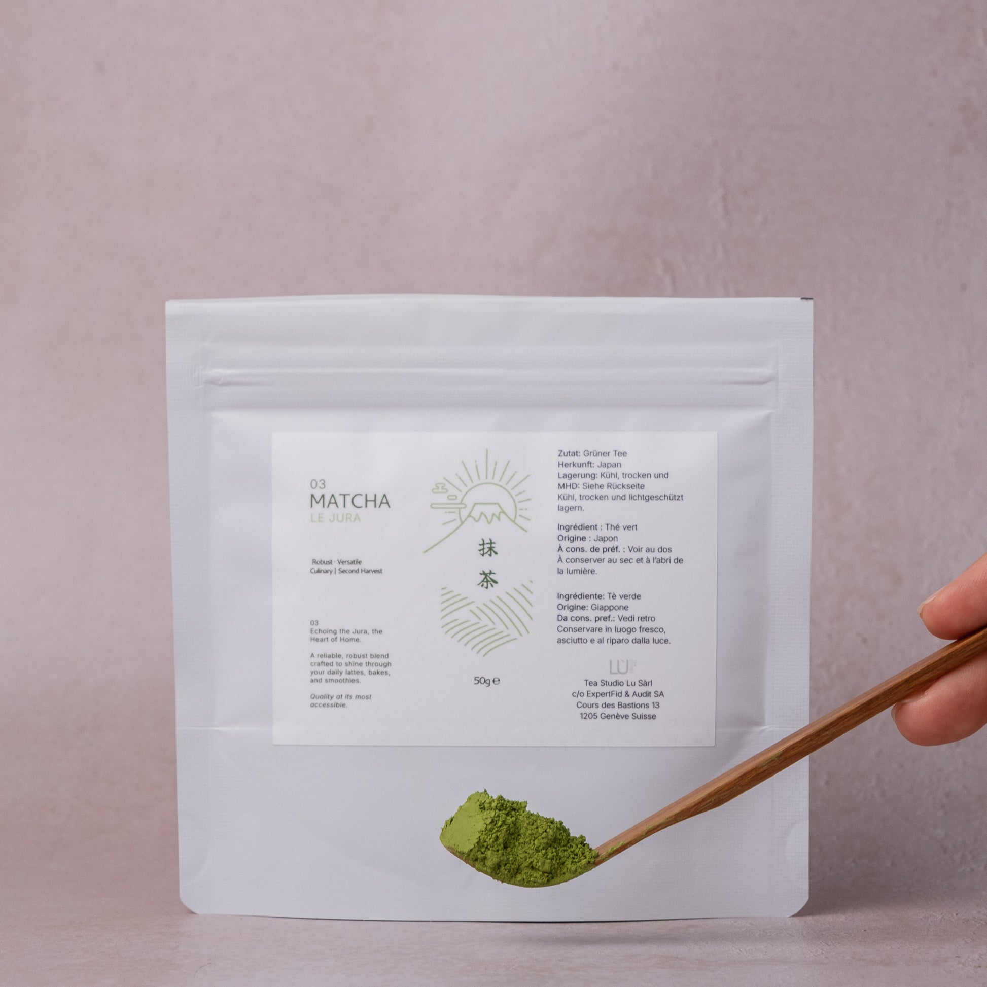 White package of matcha powder with a wooden spoonful of culinary matcha on a light pink background