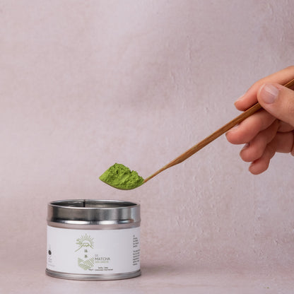 Wooden spoon lifting ceremonial matcha powder from a canister, set against a minimal light pink backdrop.