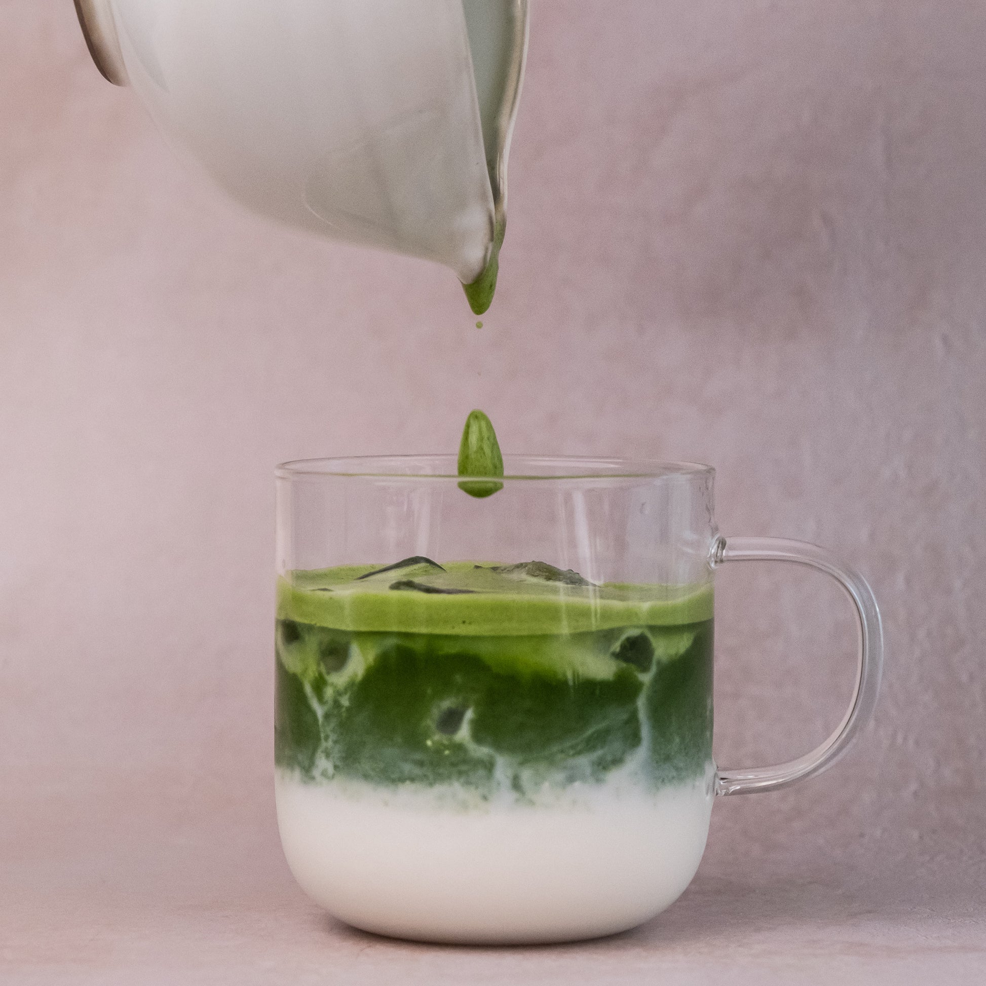 Layered iced matcha latte with milk and ceremonial matcha, pouring matcha from pitcher into glass mug on pastel pink background.
