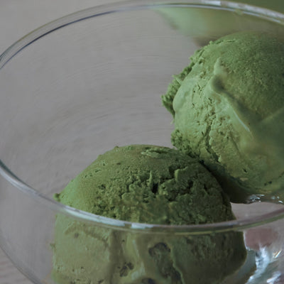 A close-up shot of two scoops of vibrant green matcha ice cream served in a clear glass bowl against a neutral background.