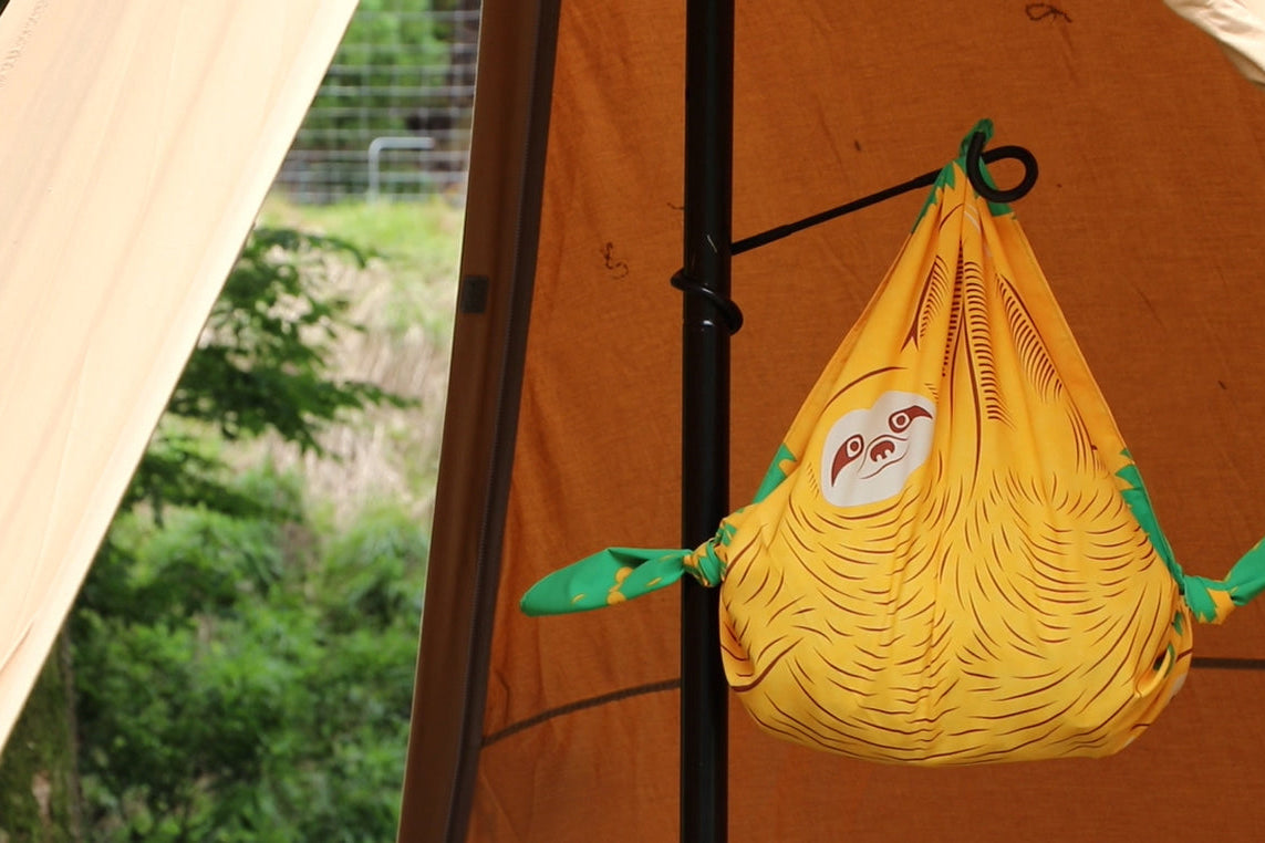 Eco-friendly outdoor camping setup featuring a yellow sloth-print Japanese furoshiki cloth tied into a hanging storage pouch inside a brown tent.